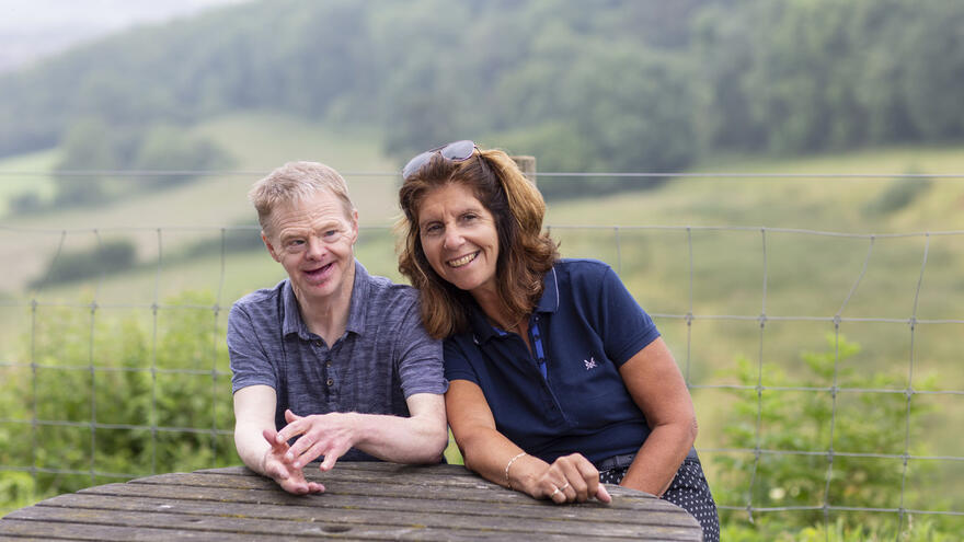 An image of a man and a woman sat next to each other at a picnic table in the countryside in summer both smiling at the camera.