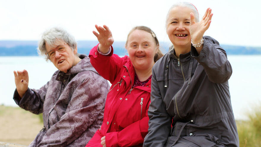 An image of three women sat on a wall, beside the beach, all waving and smiling at the camera.
